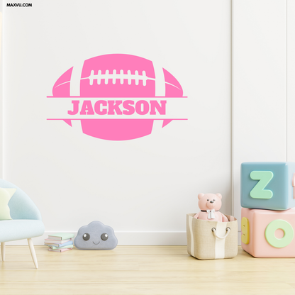 A pink football-shaped wall decal with the name 'Jackson' printed across the center, displayed on a white wall in a child's room setting with a light blue chair, a gray cloud pillow, and pastel toy blocks.